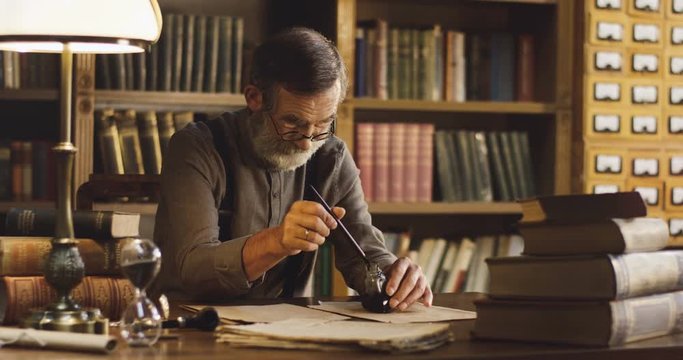 Portrait of the old Caucasian man wetting a pen in the ink and starting to write on the old wooden desk in the library room.