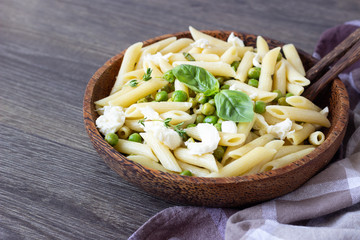 Salad with penne pasta, green peas, herbs (basil and thyme) and mozzarella cheese in a brown wooden bowl on a wooden background.