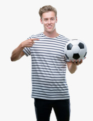 Young handsome blond man holding soccer ball very happy pointing with hand and finger