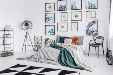 Real photo of a bed with cushions and a blanket standing in a monochromatic bedroom between a chair and a lamp and a shelf and next to a wall with botanic posters