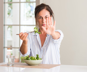 Middle aged woman eating fresh salad in a bowl at home with open hand doing stop sign with serious and confident expression, defense gesture