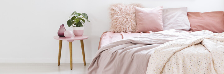 Real photo of a small table with a plant standing next to a bed with pink bedding in bedroom interior with white walls