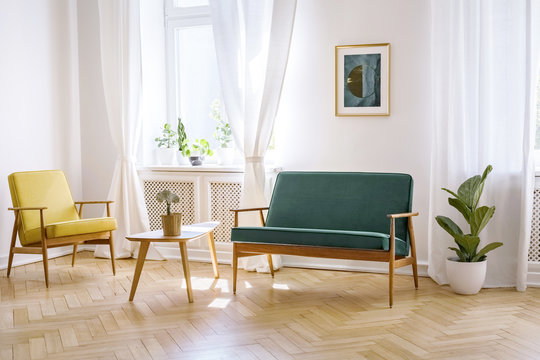 Wooden table between yellow armchair and green bench in white flat interior with poster. Real photo