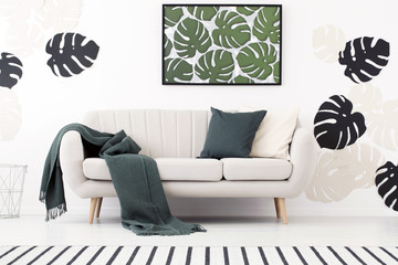 Green blanket and cushion on settee in white living room interior with poster of leaves. Real photo