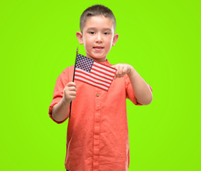 Dark haired little child holding United States flag very happy pointing with hand and finger