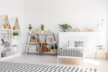 Plants and plush toy on shelves in scandi child's bedroom interior with grey sheets on bed. Real photo