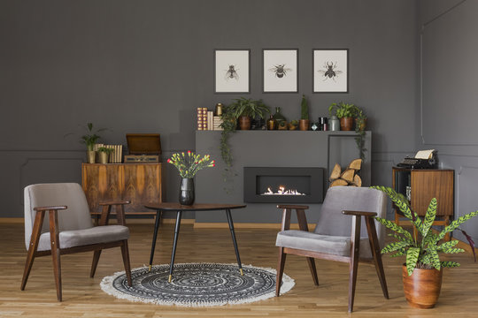 Table with flowers on rug between grey armchairs in retro living room interior with fireplace. Real photo