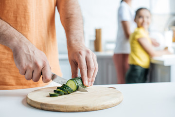 cropped shot of father cutting cucumber for salad while his wife and daughter washing dishes at kitchen