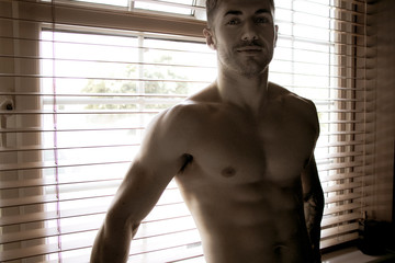 Handsome, sexy, topless, shirtless man stands in front of brown blinds and smiles at camera