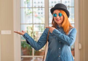 Stylish redhead woman wearing bowler hat and sunglasses amazed and smiling to the camera while presenting with hand and pointing with finger.