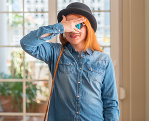 Stylish redhead woman wearing bowler hat and sunglasses peeking in shock covering face and eyes...
