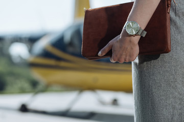 Stylish accessories. The close up of a delicate female hand with a watch on it holding a fashionable bag during the visit to a helipad
