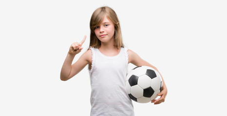 Young blonde toddler holding football ball surprised with an idea or question pointing finger with happy face, number one