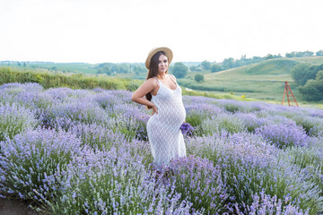 Fototapeta na wymiar attractive pregnant woman in white dress and straw hat standing at violet lavender field