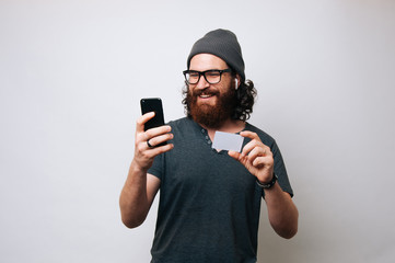 Modern young bearded man wearing a grey T-shirt and a fur hat cap. Man listening music in wireless earphones, Shopping online with credit card looking at his smartphone. Internet banking concept.