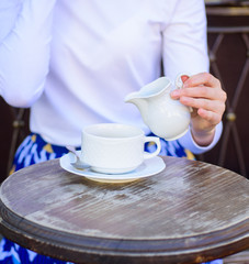 Coffee always better with milk. Hand female adding milk to black coffee outdoors cafe terrace, close up. Mug americano or black tea and jug with milk. Tips for healthier coffee drinking