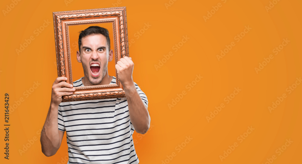 Sticker handsome young man looking through vintage art frame annoyed and frustrated shouting with anger, cra