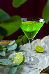A green drink with ice cubes in a glass for mojito on crumpled wrapping paper next to which lie slices of lime and a branch of green tropical leaves on a vintage wooden table.