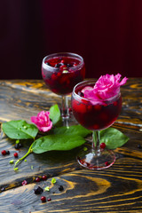 Two glasses full of red drink with berries, ice and a pink flowers, beside which lie berries and a branch of green tropical leaves on a vintage wooden table