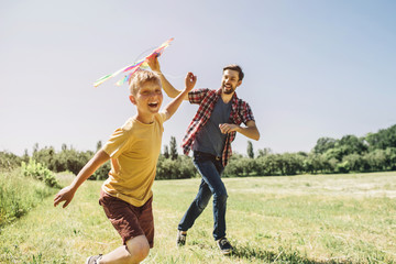 Happy kid is running on field. He is holding thread from kite. Father is holding kite. He is trying to run it to the sky. They are enjoying the moment.