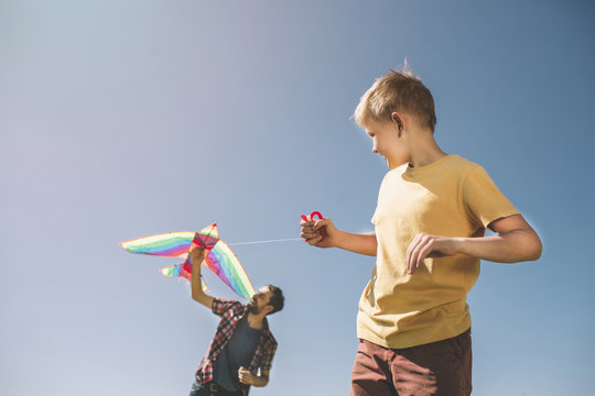 Nice picture of father and son playing with kite. Child is holding thread from it while dad is trying to run it to the sky. There is a blue background behind them.