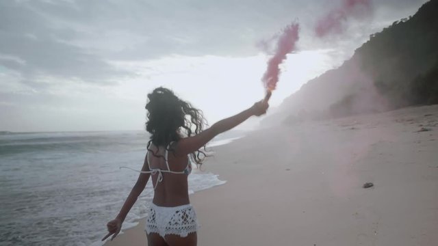 Beautiful happy girl running with colored smoke at the sandy beach during amazing sunset - video in slow motion