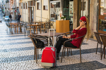 Fototapeta na wymiar Woman with red suitcase talking on the phone sitting at a table at an outdoor cafe.