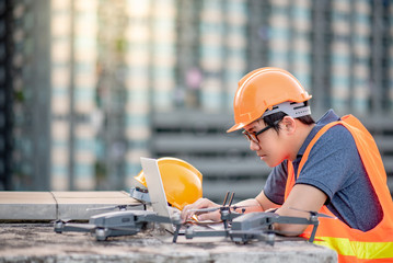 Young Asian man working with drone laptop and smartphone at construction site. Using unmanned aerial vehicle (UAV) for land and building site survey in civil engineering project.