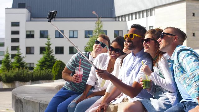 technology, friendship and international concept - group of students or friends with drinks taking picture by smartphone on selfie stick in city