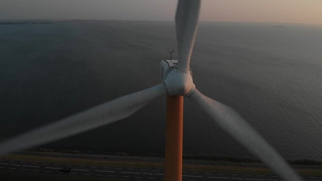 Beautiful aerial close up view of a green energy windmill at sunset, with road and car driving below and water on the background. 
