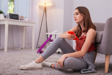 Depressive music. Sorrowful teen girl using earphones and leaning on armchair