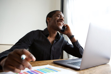 young african american business man sitting at desk with laptop talking on cellphone