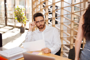 Working. Concentrated bearded man sitting at the table and holding a sheet of paper
