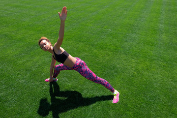 A young African-American girl in a black T-shirt, pink pants and sneakers doing sports exercises on green grass and raising her hands up. Modern sport lifestyle.