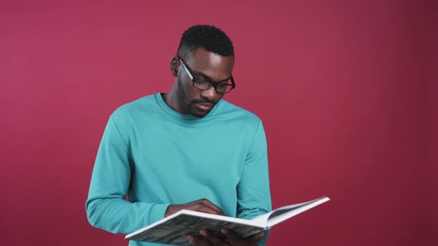 Good-looking young man in black spectacles reading book, touching pages. Cute curious African guy in mint sweater looking at camera. Pink wall.