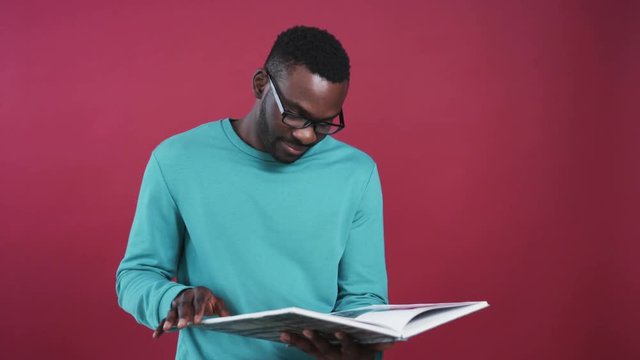 Nice young African man in glasses reading, flipping pages, looking at camera, smiling. Portrait of smart handsome guy with book. Beautiful colored wall.
