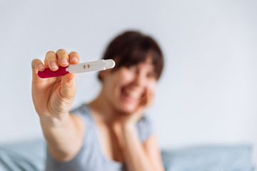 young happy woman sitting on bed looking on positive pregnancy test. blurred background