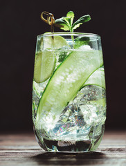 One glass with iced detox water with cucumber, lime and mint in the morning sun on a dark wood...