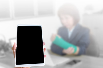 Hand holding tablet on blurred blackground injured woman with broken hand sitting and holding green cast working with laptop in office
