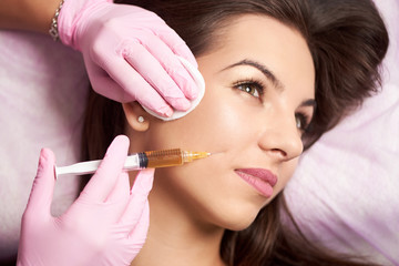 Close-up of beautiful woman getting injection in the cosmetology salon. Doctor in medical gloves with syringe injects cheeks drug.