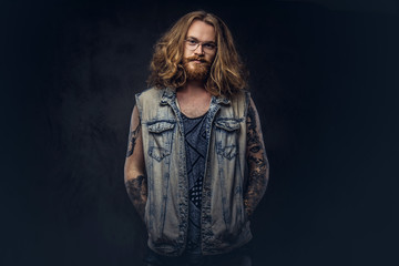 Redhead hipster male with long luxuriant hair and full beard dressed in casual clothes posing with hands in pockets in a studio, looking a camera. Isolated on a dark background.
