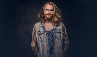 Redhead hipster male with long luxuriant hair and full beard dressed in casual clothes posing with hands in pockets in a studio, looking a camera. Isolated on a dark background.