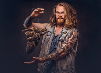 Portrait of a tattoed redhead hipster male with long luxuriant hair and full beard dressed in a t-shirt and jacket holds a keeps the scarecrow of an owl in a studio. Isolated on the dark background.