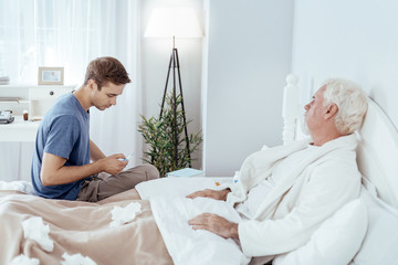 High body temperature. Senior man lying in bed and concerned man holding thermometer