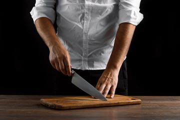 Hands of the chef close-up with a knife and a wooden board on a dark background. The concept of cooking, cooking, recipe dishes.