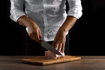 The chef cuts a piece of fresh meat on a wooden board against a dark background, hands close-up. The concept of cooking, cooking meat, a recipe for meat dishes.