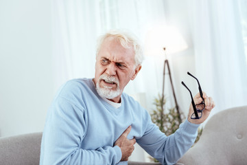 Heart rhythm disturbances. Painful senior man touching chest and holding glasses