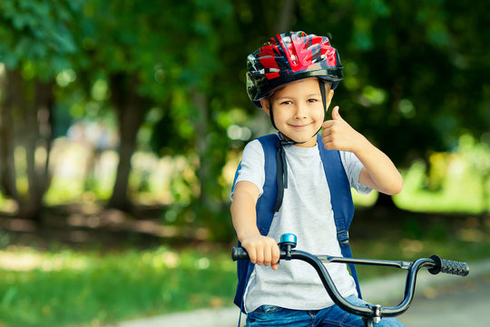 Little boy learns to ride a bike in the park near the home. Kid shows the thumbs up on bicycle. Happy smiling child in helmet riding a cycling.