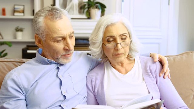 Close up portrait of grandparents reading boook on the sofa at home in the living room while their grandson and granddaughter running to the room and hugging them.
