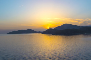 Mediterranean landscape. Sunset on the sea with flowing bright colored rays of the sun through the clouds. Silhouettes of mountains. Silhouettes the ship. Background.